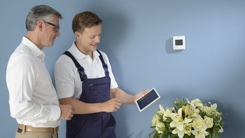 In einem Raum stehen zwei Männer, der Linke ist ein Kunde und der Rechte ein Mechatroniker, der hält ein Tablet in der Hand. Beide schauen zum Tablet. An der Wand neben den Männern, ist eine Klima Steuerung montiert.